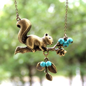 May include: A gold-tone necklace features a squirrel figurine perched on a branch with turquoise blue berries. The squirrel is facing left and has a bushy tail. The necklace is perfect for nature lovers.