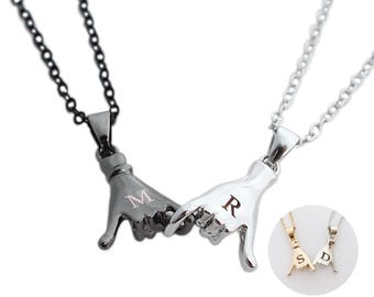 Peut inclure: Deux colliers avec des pendentifs en forme de mains faisant un geste de promesse. Un pendentif est noir avec la lettre M, l'autre est argenté avec la lettre R. Un encart circulaire montre des versions dorées et argentées avec les lettres S et D.