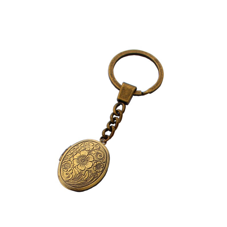 Puede incluir: Un llavero dorado con un dije ovalado adornado con un dise&ntilde;o floral. La cadena est&aacute; unida a un anillo circular.
