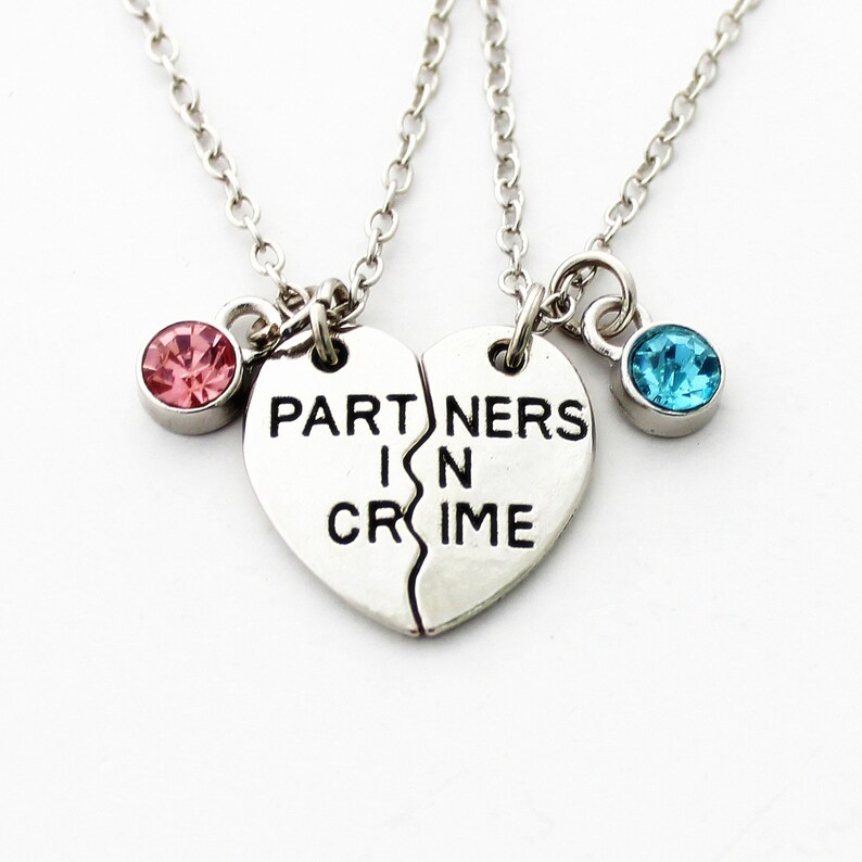 May include: Silver-toned necklace set featuring two halves of a heart that, when joined, read "PARTNERS IN CRIME." One half has a pink gemstone, the other a blue gemstone. The necklaces are connected by delicate chains.