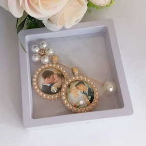 May include: A gold-toned brooch featuring two circular photo pendants framed by small white pearls. One pendant shows a couple embracing, and the other shows a couple kissing. A flower-shaped accent with white pearls sits at the top of the pin.