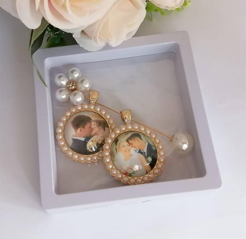 May include: A gold-toned brooch featuring two circular photo pendants framed by small white pearls. One pendant shows a couple embracing, and the other shows a couple kissing. A flower-shaped accent with white pearls sits at the top of the pin.