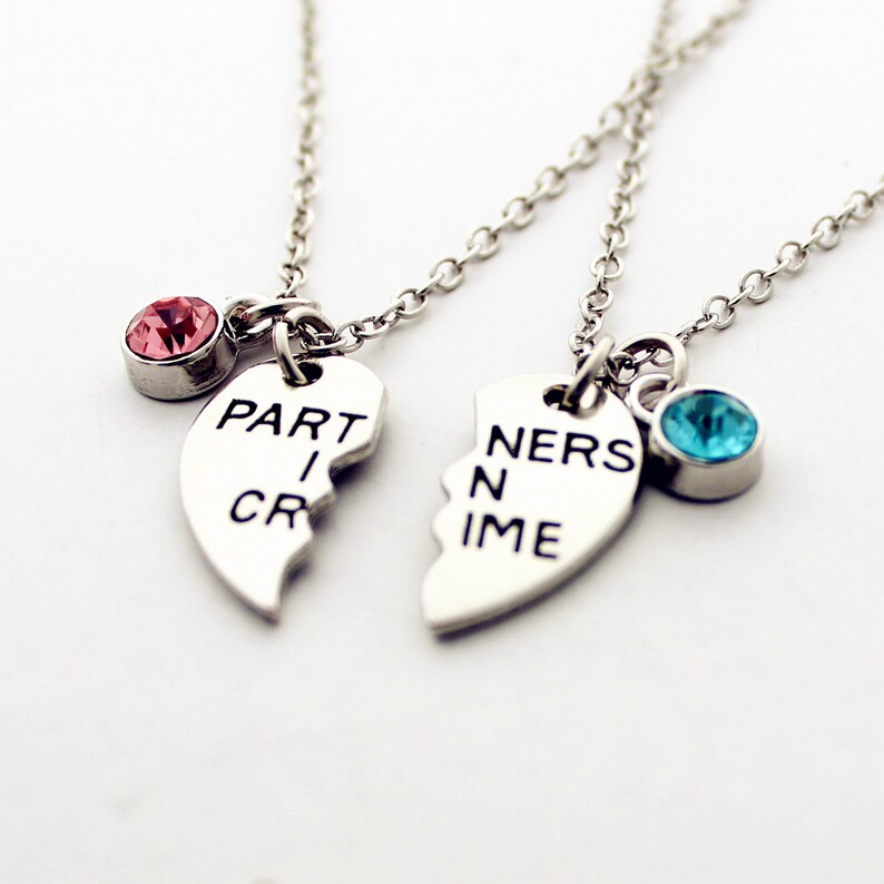 May include: Two silver necklaces with heart-shaped pendants that read "PART I CRIME" and "NERS N IME". Each pendant is connected to a chain and has a colored gemstone. One has a pink gem, the other a blue gem.