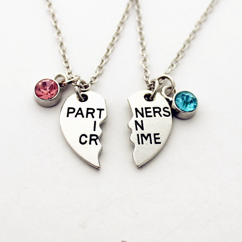 May include: Two silver necklaces, each with a half-heart pendant that reads "PART I CRIME" and "NERS IN TIME". One necklace has a pink gemstone, the other a blue gemstone. The necklaces are on a white background.