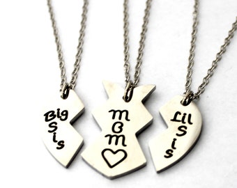 May include: Three silver necklaces with heart-shaped pendants. The pendants are broken into three pieces, each with a different inscription: "Big Sis", "Mom", and "Lil Sis".