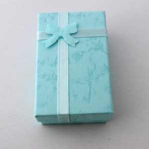 May include: A rectangular, light blue gift box with a textured surface. A light blue ribbon with a bow is wrapped around the box. The box is closed and ready for gifting.