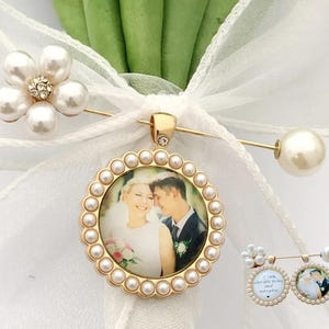May include: A gold-toned brooch featuring a circular frame bordered by small white pearls, enclosing a photograph of a couple. The brooch is attached to a white ribbon and includes a flower-shaped accent with pearls and clear crystals.