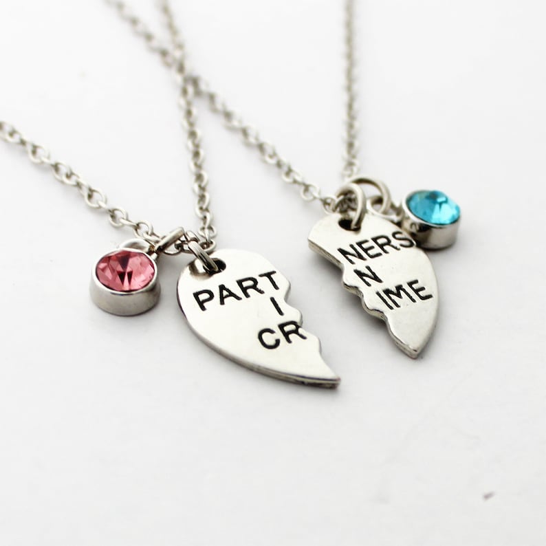 May include: Two silver necklaces with heart-shaped pendants, each split in half. One pendant reads "PART I CR" and has a pink gemstone. The other reads "NERS N IME" and has a blue gemstone. The necklaces are connected by delicate chains.
