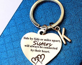 May include: Silver heart-shaped keychain with the text "Side by Side or miles apart, Sisters will always be connected by their heart." and two intertwined hearts. The keychain has a silver ring and a silver letter K charm.