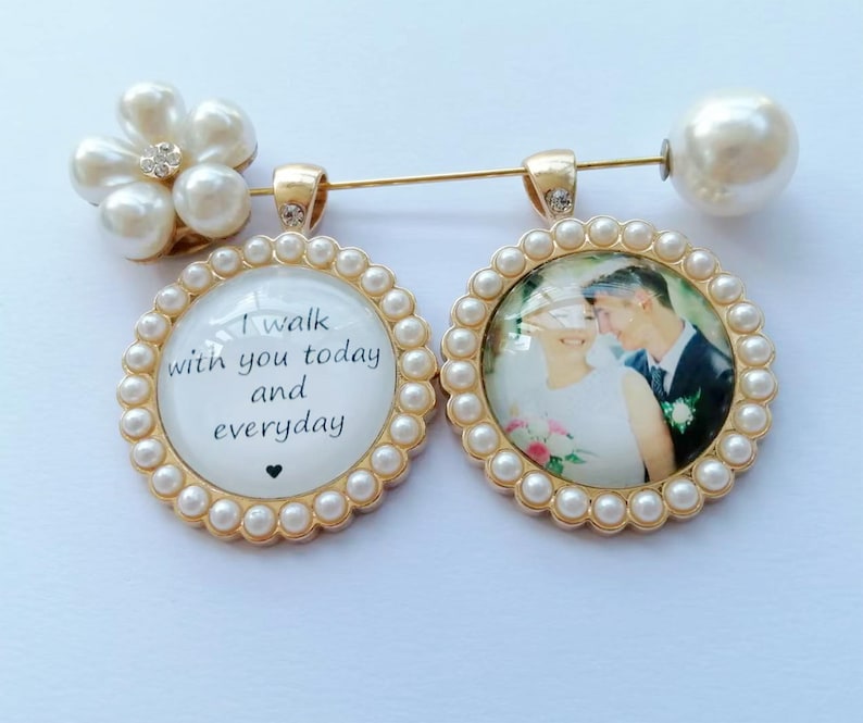 May include: A gold-toned brooch featuring two circular pendants framed by small white faux pearls. One pendant displays the text I walk with you today and everyday with a small heart. The other pendant shows a photograph of a couple. A large faux pearl sits at one end of the pin.