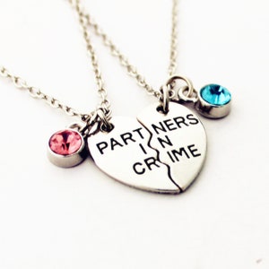 May include: Two silver necklaces with a heart-shaped pendant that reads "PARTNERS IN CRIME". One necklace has a pink gemstone, the other a blue gemstone. The necklaces are on a white background.