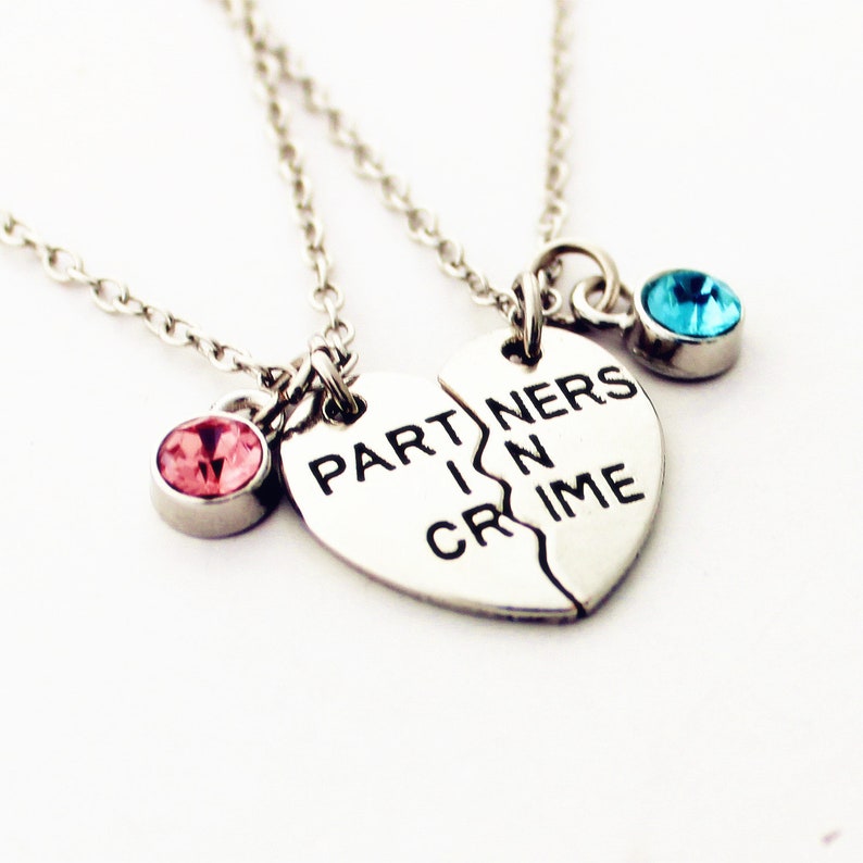 May include: Two silver necklaces with a heart-shaped pendant that reads "PARTNERS IN CRIME". One necklace has a pink gemstone, the other a blue gemstone. The necklaces are on a white background.