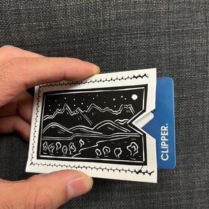 May include: A black and white landscape design on a white card holder. The card holder is open, revealing a blue card with the word "CLIPPER." The design features mountains, trees, and a starry sky.