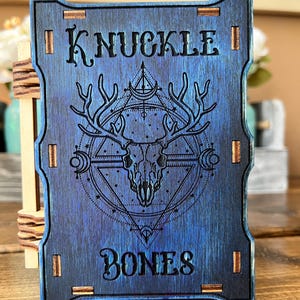 May include: A blue wooden box with the words "KNUCKLE BONES" engraved on the front. The box features a black and white design of a skull with antlers inside a geometric pattern.