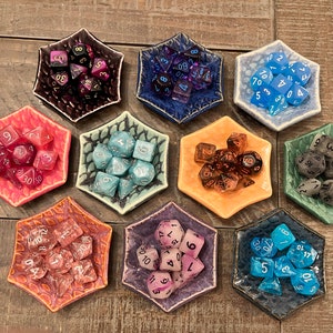 Könnte beinhalten: Neun sechseckige Keramikschalen mit strukturierter Oberfläche, jede mit einem Satz bunter Würfel. Die Würfel sind in verschiedenen Farben, darunter Lila, Blau, Grün, Orange und Schwarz.
