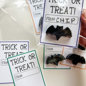 K&ouml;nnte beinhalten: Schwarze fledermausf&ouml;rmige Halloween-Gastgeschenke mit "Trick or Treat!" Karten. Die Karten, in durchsichtigen Beuteln, haben "From: Chip" Etiketten. Andere Karten haben leere "From:" Etiketten und verschiedenfarbige R&auml;nder.