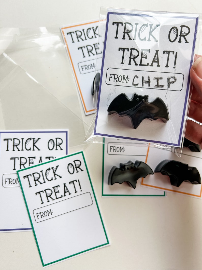 K&ouml;nnte beinhalten: Schwarze fledermausf&ouml;rmige Halloween-Gastgeschenke mit "Trick or Treat!" Karten. Die Karten, in durchsichtigen Beuteln, haben "From: Chip" Etiketten. Andere Karten haben leere "From:" Etiketten und verschiedenfarbige R&auml;nder.