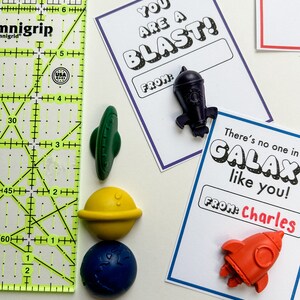 May include: A collection of space-themed Valentine's Day cards and wax crayon shapes. The cards have phrases like "You are a Blast!" and "There's no one in GALAX like you!" with a name field. The crayons are shaped like a rocket, planet, and UFO.