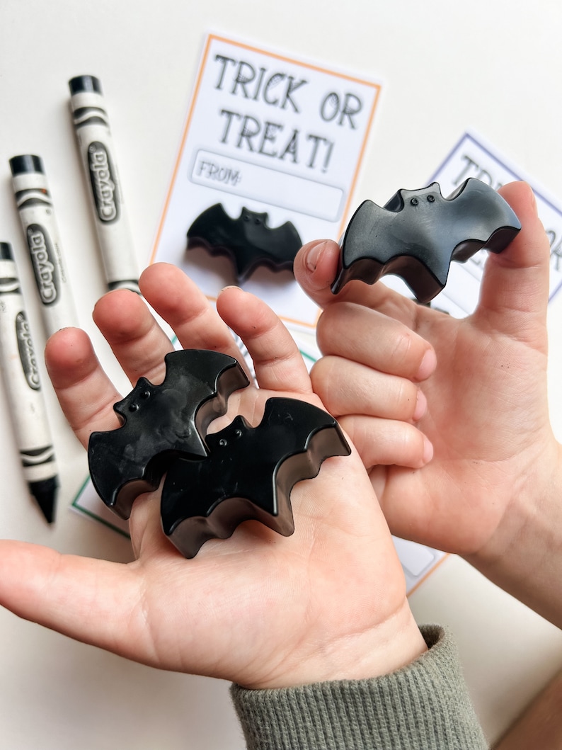 K&ouml;nnte beinhalten: Schwarze, fledermausf&ouml;rmige Wachsmalstifte werden in den H&auml;nden einer Person gehalten. Wei&szlig;e Crayola-Stifte und eine "Trick or Treat!"-Karte sind im Hintergrund zu sehen. Die Karte hat ein Feld f&uuml;r "From:"