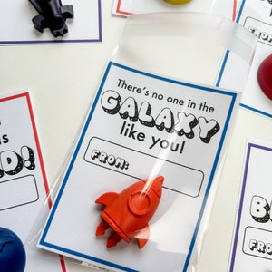 May include: A collection of small, colorful crayon shapes, including a red rocket, a yellow helmet, and a blue globe, are displayed on white cards. Each card has a blue or red border and includes the text "There's no one in the GALAXY like you!" and a "FROM:" section.
