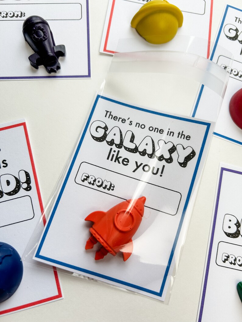 May include: A collection of small, colorful crayon shapes, including a red rocket, a yellow helmet, and a blue globe, are displayed on white cards. Each card has a blue or red border and includes the text "There's no one in the GALAXY like you!" and a "FROM:" section.