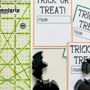 K&ouml;nnte beinhalten: Halloween-Artikel. Schwarze fledermausf&ouml;rmige Dekorationen und wei&szlig;e Karten mit orangefarbenen R&auml;ndern mit der Aufschrift "TRICK OR TREAT!" und "FROM:" werden neben einem gelb-gr&uuml;nen Lineal angezeigt.