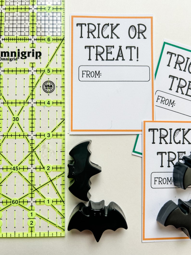 K&ouml;nnte beinhalten: Halloween-Artikel. Schwarze fledermausf&ouml;rmige Dekorationen und wei&szlig;e Karten mit orangefarbenen R&auml;ndern mit der Aufschrift "TRICK OR TREAT!" und "FROM:" werden neben einem gelb-gr&uuml;nen Lineal angezeigt.