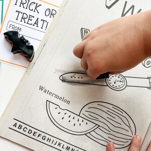 K&ouml;nnte beinhalten: Eine Kinderhand malt eine schwarz-wei&szlig;e Illustration einer Wassermelone aus. Die Seite zeigt auch den Buchstaben "W" und das Wort "Watermelon". Zwei schwarze Fledermaus-Wachsmalstifte und eine "Trick or Treat"-Karte sind ebenfalls zu sehen.