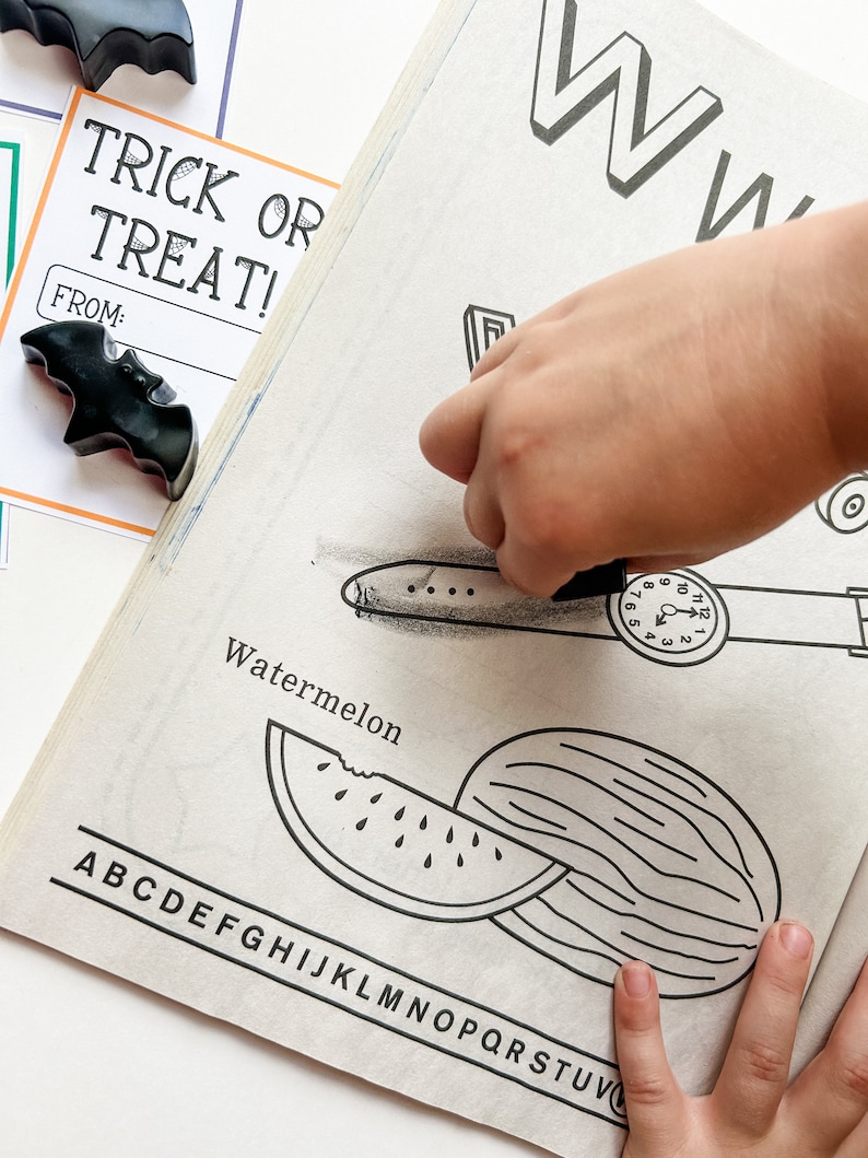 K&ouml;nnte beinhalten: Eine Kinderhand malt eine schwarz-wei&szlig;e Illustration einer Wassermelone aus. Die Seite zeigt auch den Buchstaben "W" und das Wort "Watermelon". Zwei schwarze Fledermaus-Wachsmalstifte und eine "Trick or Treat"-Karte sind ebenfalls zu sehen.