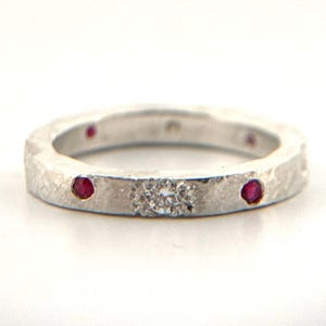 May include: A silver ring with a textured surface. The ring features three small, round, red gemstones and a cluster of small, clear gemstones. The ring is set against a white background.