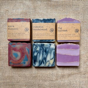 May include: Three rectangular shea butter bar soaps, each 4 oz (113g). The first is red and blue marbled, labeled 'warm flannel'. The second is black and white marbled, labeled 'pumpkin sandalwood'. The third is purple with gold lines, labeled 'fig harvest'.