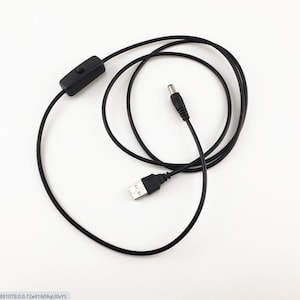 Puede incluir: Cable de alimentación negro con un conector USB, un conector de alimentación de CC y un interruptor de encendido/apagado en línea. El cable está enrollado y sobre una superficie blanca. Los conectores son negros.