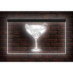 May include: A neon sign featuring a cocktail glass design with the words "BAR OPEN" in white. The sign is set against a dark brick wall and framed with a white border. A lemon slice and a straw are included in the cocktail glass design.