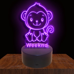 Puede incluir: Una luz de neón morada con forma de mono de dibujos animados con la palabra "Weeknd" debajo. La luz está sobre una base negra.