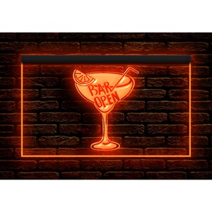 May include: An orange illuminated sign featuring a cocktail glass design with the words "BAR OPEN". The sign has a black border and is set against a dark brick wall. The cocktail glass includes a citrus slice and a straw.