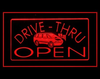May include: Red neon sign with the words "Drive-Thru Open" and a cartoon car. The sign is outlined in red and has a black background.