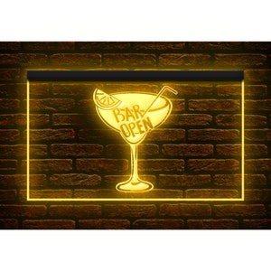 May include: A neon sign with a yellow glow, featuring a cocktail glass design with a lemon slice and a straw. The words "BAR OPEN" are inside the glass. The sign is set against a brown brick wall.