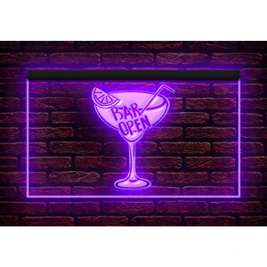 May include: A neon sign with a purple glow against a brick wall. The sign is shaped like a cocktail glass with a lemon slice and a straw. The words "BAR OPEN" are written inside the glass.