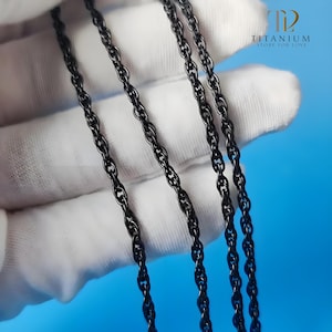 Puede incluir: Primer plano de varios collares de cadena negros. Las cadenas están hechas de pequeños eslabones entrelazados. El fondo es de color azul claro. El texto "TITANIUM STORE FOR LOVE" es visible en la esquina superior derecha.