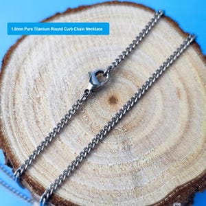 May include: A close-up of a 1.8mm pure titanium round curb chain necklace. The silver-colored chain is displayed on a wooden surface with a blue background. The necklace is fastened with a clasp.
