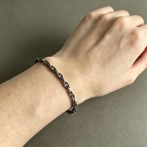 May include: A dark silver-toned metal chain bracelet featuring a series of interconnected oval links. The bracelet has a polished finish and rests securely around the wrist, showcasing a simple and uniform design with consistent link sizing throughout.
