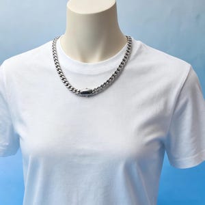 Pode incluir: Um colar de corrente de elos prateado com fecho retangular é exibido em uma camiseta branca. O colar é feito de elos grossos e interligados. O fundo é um gradiente de azul claro.