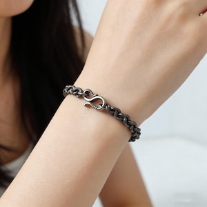May include: A dark gray bracelet with a woven rope design. The bracelet features a hook clasp closure. The jewelry is worn on a wrist, showcasing the intricate texture and style of the accessory.