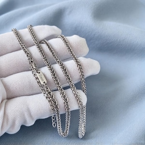 May include: A silver-toned wheat chain necklace featuring a metallic tag engraved with the word titanium. The chain has a woven, textured link design and includes an adjustable extender section with a lobster claw clasp closure.