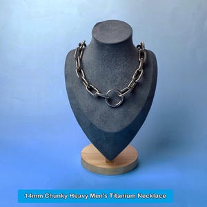 Puede incluir: Un collar de titanio grueso con eslabones de 14 mm y un colgante circular grande. El collar se exhibe sobre un busto gris sobre un fondo azul. El texto en la parte inferior dice "14mm Chunky Heavy Men's Titanium Necklace."