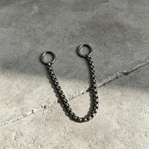 May include: A silver-toned chain with two circular rings at each end. The chain is composed of small, rectangular links. The piece is displayed on a gray concrete surface, with natural light casting shadows.