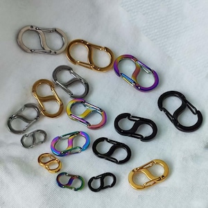 Puede incluir: Una colección de mosquetones de resorte metálicos en plata, oro, negro y colores arcoíris iridiscentes. Los ganchos, de forma ovalada, presentan un diseño de doble lazo, adecuados para llaveros, bolsos u otros accesorios.