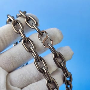 May include: Close-up of two silver metal chains with oval links and a silver clasp. The chains are held against a blue background. The chains are made of a shiny, metallic material.