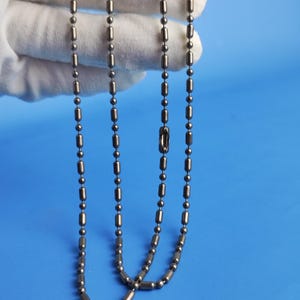 May include: Close-up of a gunmetal-colored ball chain necklace. The necklace is composed of small, round beads and elongated, cylindrical links. The chain is displayed against a blue background.