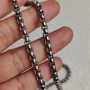 May include: A silver chain necklace with a box chain design. The chain is shown on a hand.